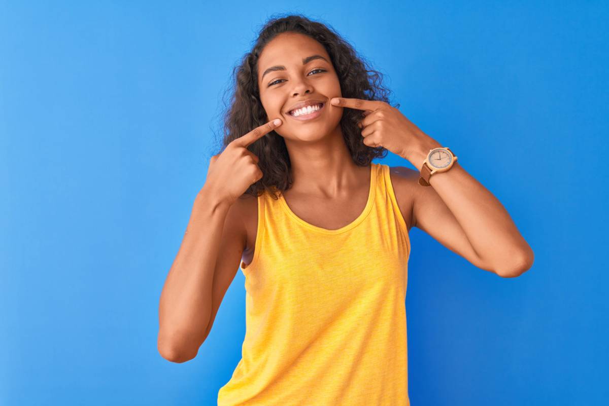 A woman in a yellow tank top smiles while playfully pointing her finger to her mouth, suggesting a secret or surprise.