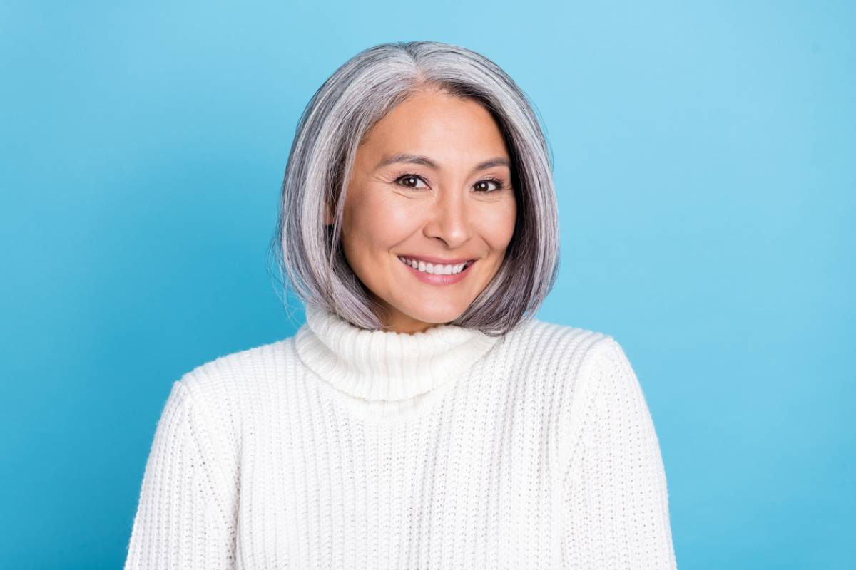 A smiling woman with gray hair is wearing a cozy white sweater, radiating warmth and positivity.