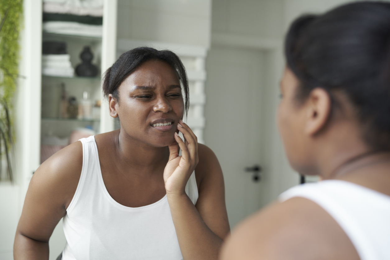 The image shows an African American woman holding her jaw in pain due to tooth grinding. The image shows that there are long-term effects of teeth grinding.